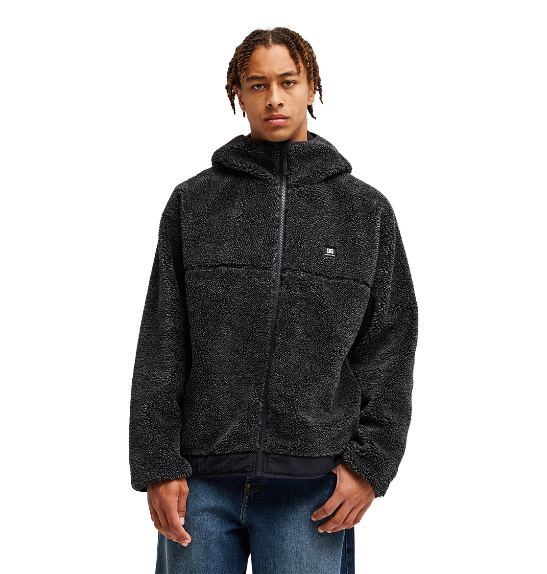 A person wearing the DC Shoes Coldstag Sherpa Reversible textured black zip-up hoodie with a hood—designed for cold-weather comfort—stands against a plain white background. They have short, braided hair and are dressed in blue jeans.