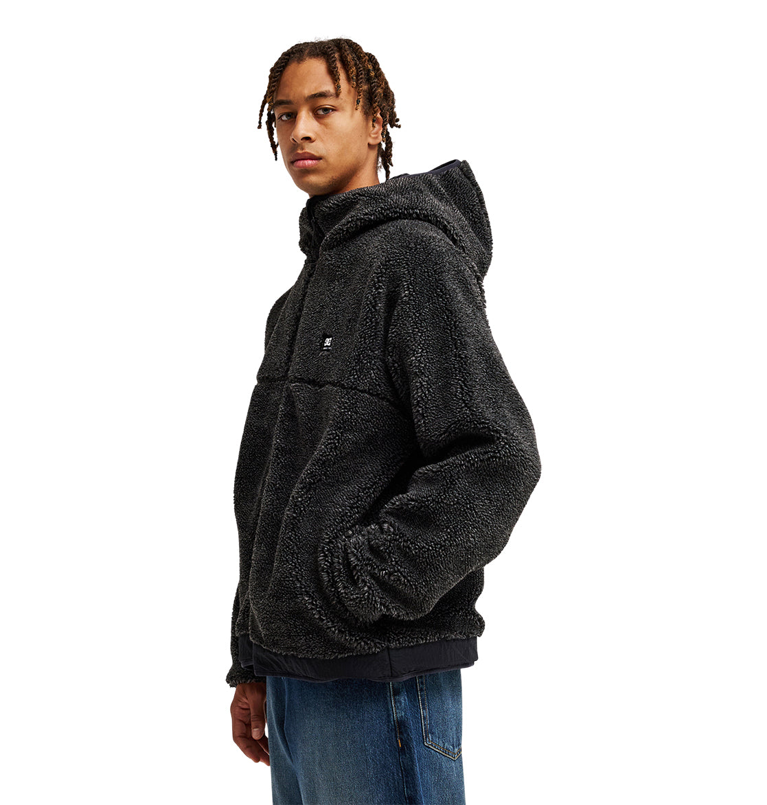 A person wearing the DC Shoes Coldstag Sherpa Reversible jacket—a black, fuzzy fleece design with a hood—paired with blue jeans stands slightly turned against a white background, highlighting its cozy cold-weather comfort.