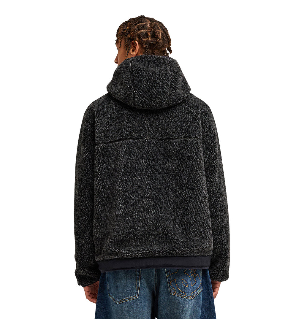 A person with braided hair is pictured from behind, wearing the DC Shoes Coldstag Sherpa Reversible fleece jacket in black for cozy cold-weather comfort, paired with blue jeans featuring patchwork designs on the pockets.