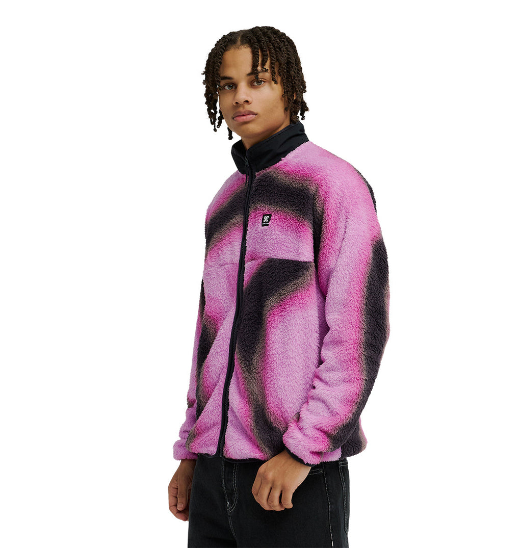 A person with braided hair wears the DC Shoes Lynx Fleece—a soft, fuzzy pink and black zip-up jacket with a black collar—paired with black pants, standing against a plain white background.