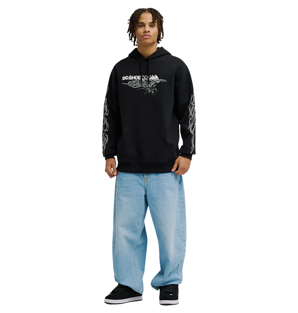 A person stands facing forward, wearing the Men's Side Flame Hoodie from DC Shoes—a black pullover featuring bold DC graphics—paired with light blue baggy jeans. Their shoulder-length hair frames their face, and they wear black shoes. The cozy fleece warmth of the hoodie adds comfort, while the background is plain white.