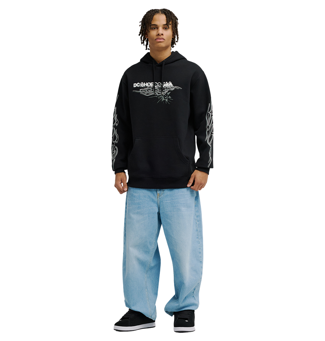 A person stands facing forward, wearing the Men's Side Flame Hoodie from DC Shoes—a black pullover featuring bold DC graphics—paired with light blue baggy jeans. Their shoulder-length hair frames their face, and they wear black shoes. The cozy fleece warmth of the hoodie adds comfort, while the background is plain white.