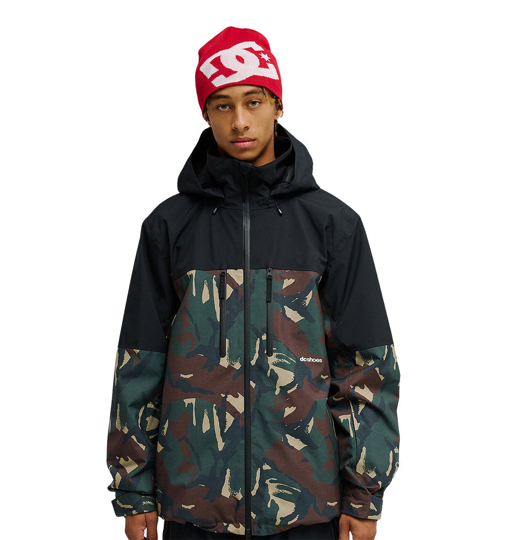 A person wearing a red DC Shoes beanie and a black and camouflage DC Shoes Edge Jacket 20K with a front zipper, featuring advanced waterproofing for superior snow performance, stands against a plain white background.