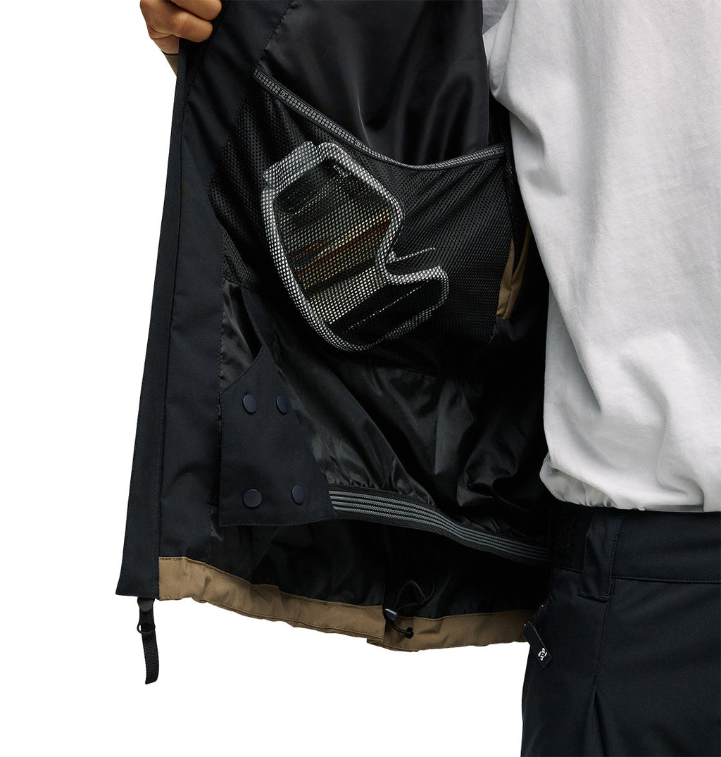 A person opens the Edge Jacket 20K by DC Shoes, showcasing its black, breathable design and revealing a mesh inner pocket that holds a smartphone. The person pairs the jacket with dark pants and a white shirt.