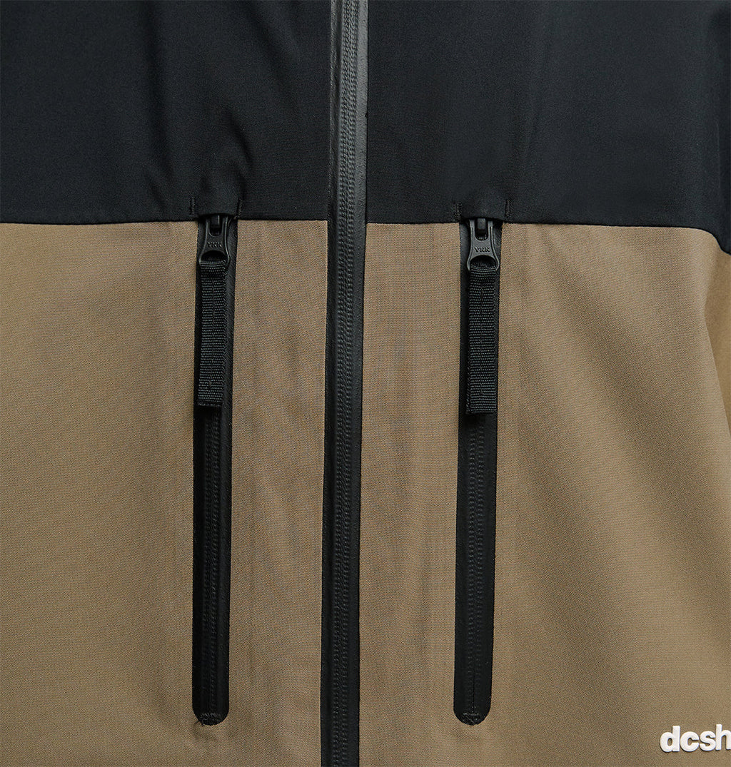 Close-up of the DC Shoes Edge Jacket 20K, showcasing its two-tone design with a black upper section and brown lower section. The jacket features a black waterproof zipper and two vertical zippered pockets for breathable protection, with the dcsh logo partially visible in the bottom right corner.