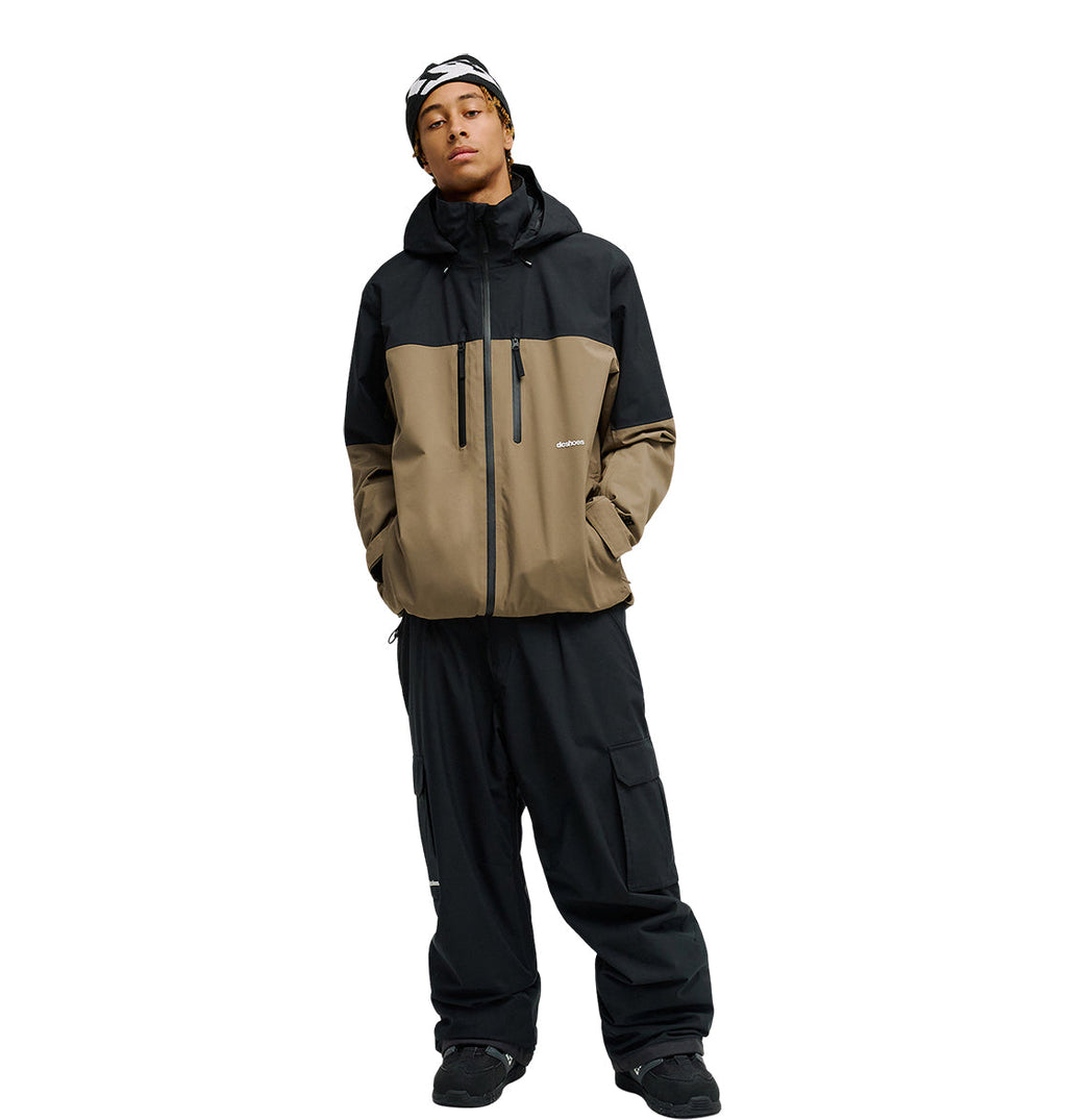 A person stands against a plain white background wearing the DC Shoes Edge Jacket 20K—a two-tone black and brown waterproof jacket—paired with black cargo pants, black shoes, and a black and white beanie.