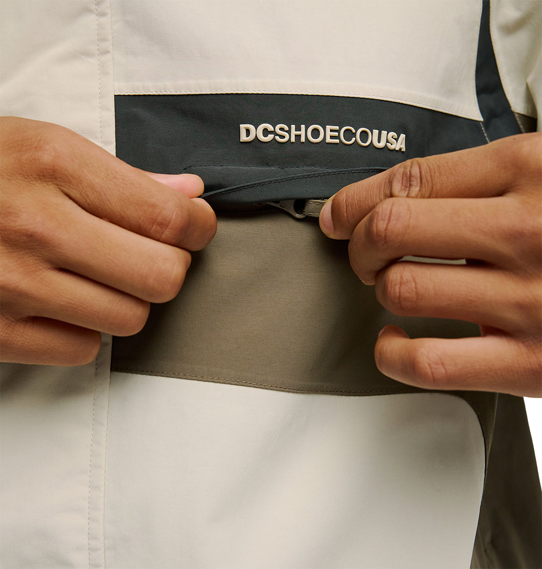Close-up of hands opening a zippered pocket on the color-blocked Mountain Lab Jacket 20K by DC Shoes, featuring “DCSHOECOUSA” above the pocket and showcasing its breathable protection and dependable snow performance.
