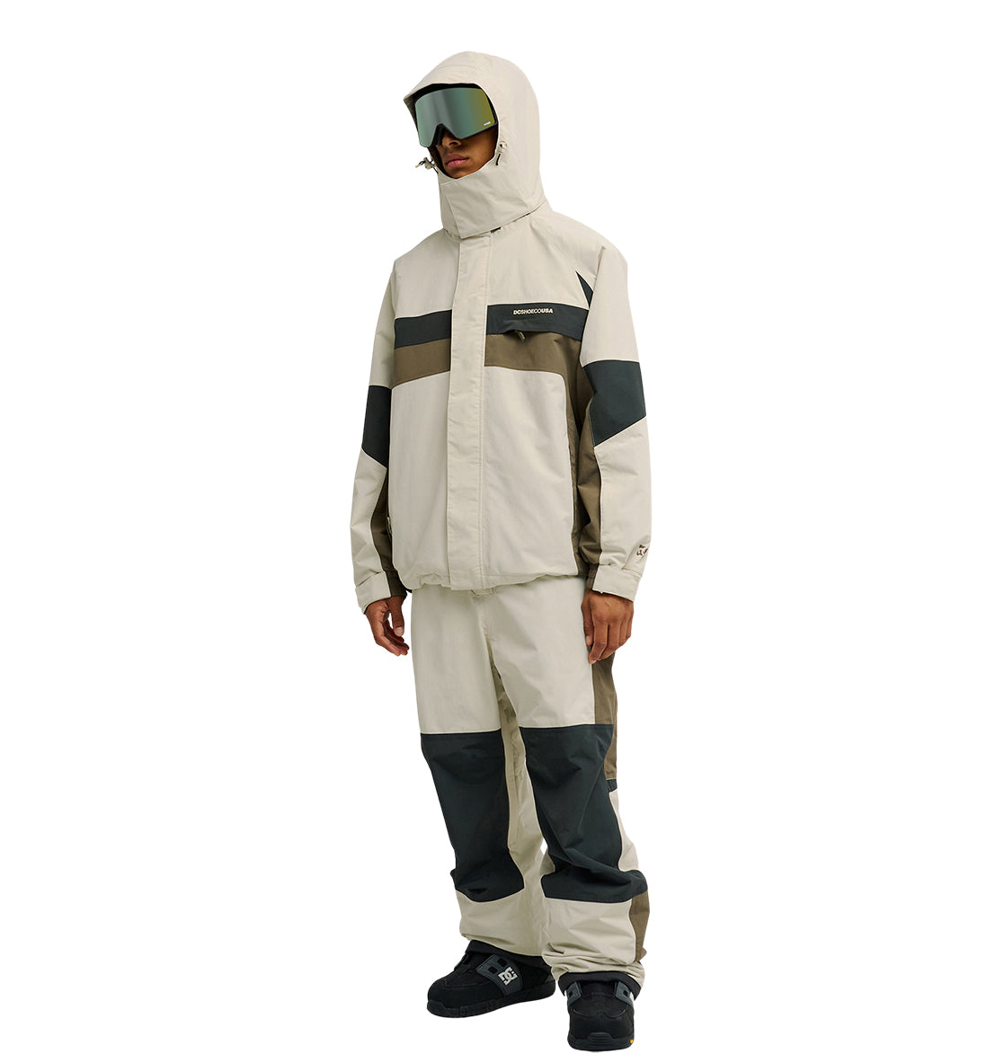 A person is wearing the DC Shoes Mountain Lab Jacket 20K, a beige and gray hooded winter jacket designed for breathable protection and enhanced snow performance, paired with matching pants, black boots, and large reflective ski goggles, standing against a white background.