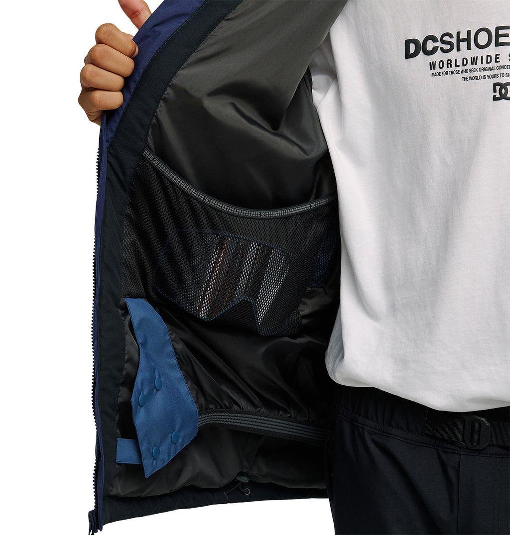 A person opens the DC Shoes Coldsmoke Jacket 10K to reveal its black interior pockets and blue lining details, highlighting its breathable protection. The individual is also wearing a white DC Shoes t-shirt and dark pants.