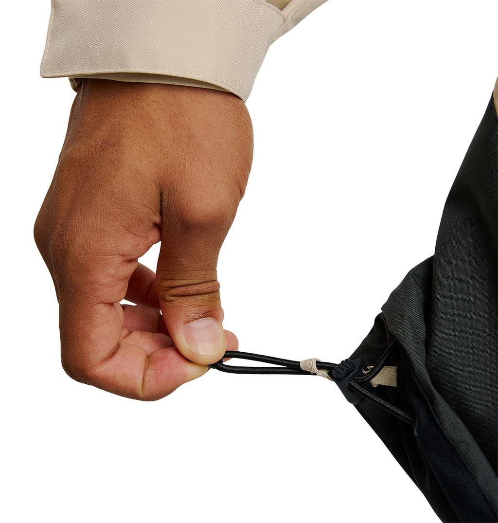 A hand pulls the adjustable drawstring on a DC Shoes Coldsmoke Jacket 10K made of dark, insulated fabric, cinching it tighter. The person is wearing a light-colored long sleeve shirt underneath.