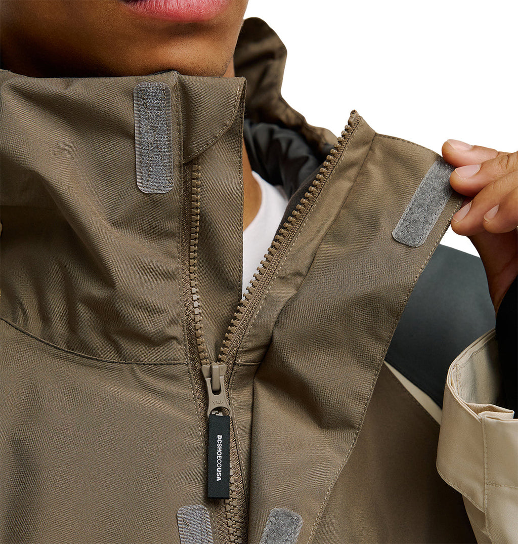 A person zips up the DC Shoes Coldsmoke Jacket 10K in brown, featuring gray velcro patches on the collar that emphasize its secure fastening and breathable protection. Only the lower half of the face and a hand are visible.