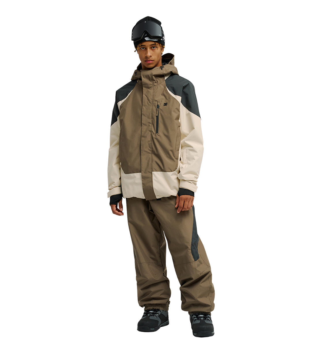 A person wearing the DC Shoes Coldsmoke Jacket 10K in brown and beige—designed for excellent snow performance—paired with matching ski pants, black gloves, black boots, and a black ski helmet with goggles, standing against a plain white background.
