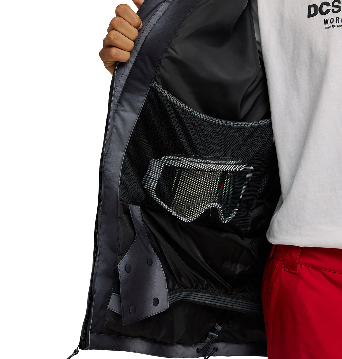 A person opens a black DC Shoes Basis Jacket 10K, revealing a mesh inner pocket holding a pair of goggles. The person wears a white t-shirt and red pants.