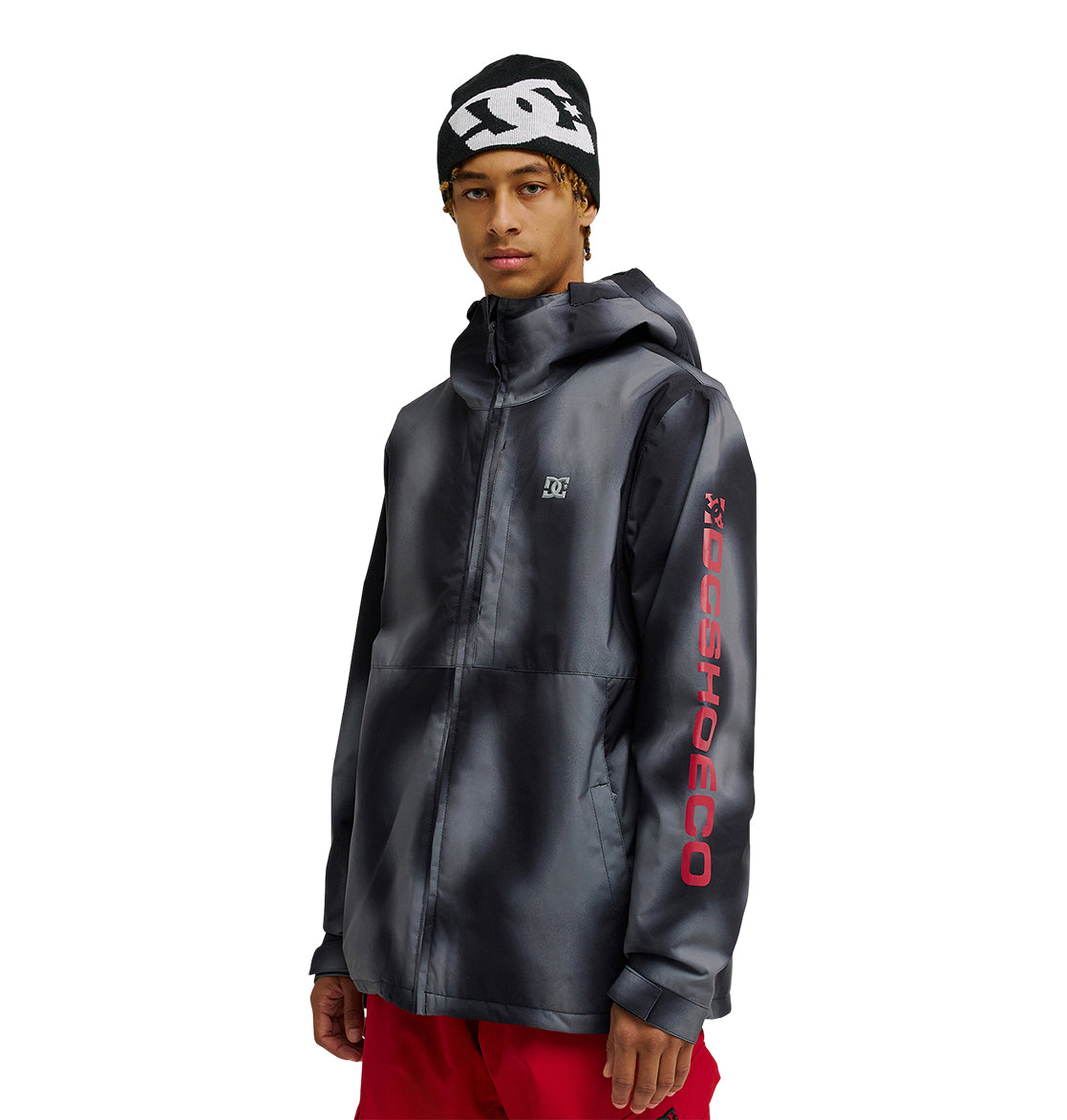 A person wearing a black DC Shoes Basis Jacket 10K with a hood, featuring a red DC SHOES logo on the sleeve, red pants, and a black beanie with a white DC Shoes logo, standing against a white background.