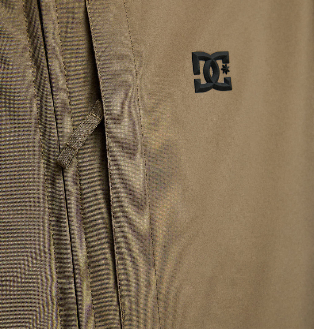 Close-up of the tan DC Shoes Basis Jacket 10K, showcasing a black brand logo on the right and a vertical zippered pocket seam on the left, emphasizing its insulated and breathable design.