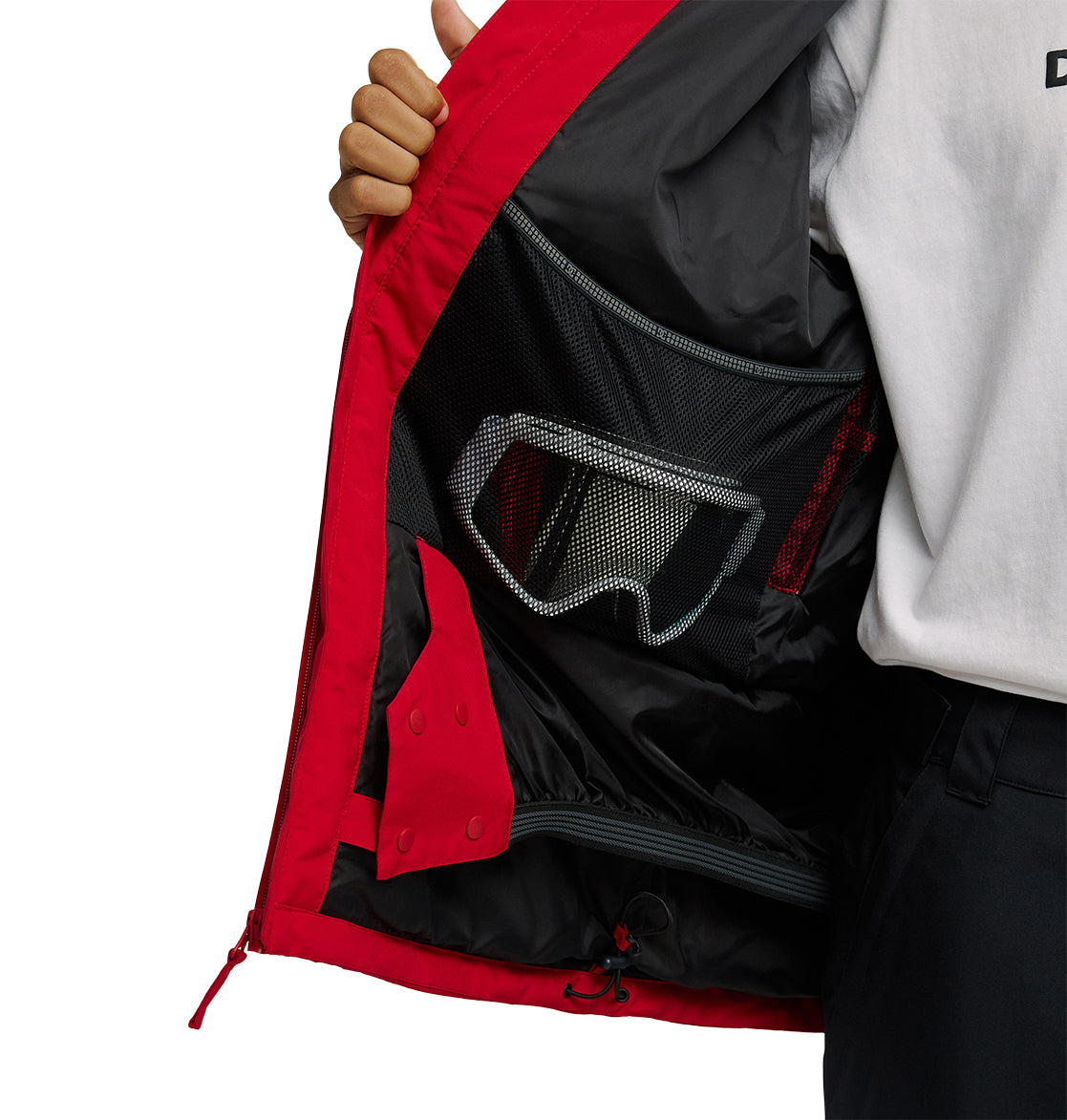 A person holds open a red DC Shoes Basis Jacket 10K, revealing the inside pocket with a mesh section containing ski goggles.