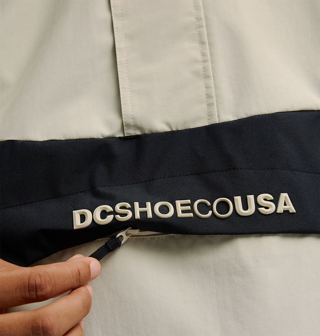 A hand unzips a black pocket on the light-colored DC Shoes Mountain Lab Anorak 10K jacket, providing easy access while ensuring breathable protection. The word DCSHOECOUSA is printed in large white letters above the zipper.