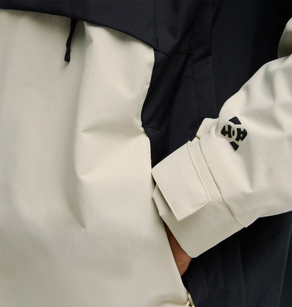 A person wearing the DC Shoes Mountain Lab Anorak 10K in a two-tone black and off-white design with one hand in the pocket. The sleeve features a small black HOKA logo near the wrist, highlighting the jacket’s breathable protection for all-day comfort.