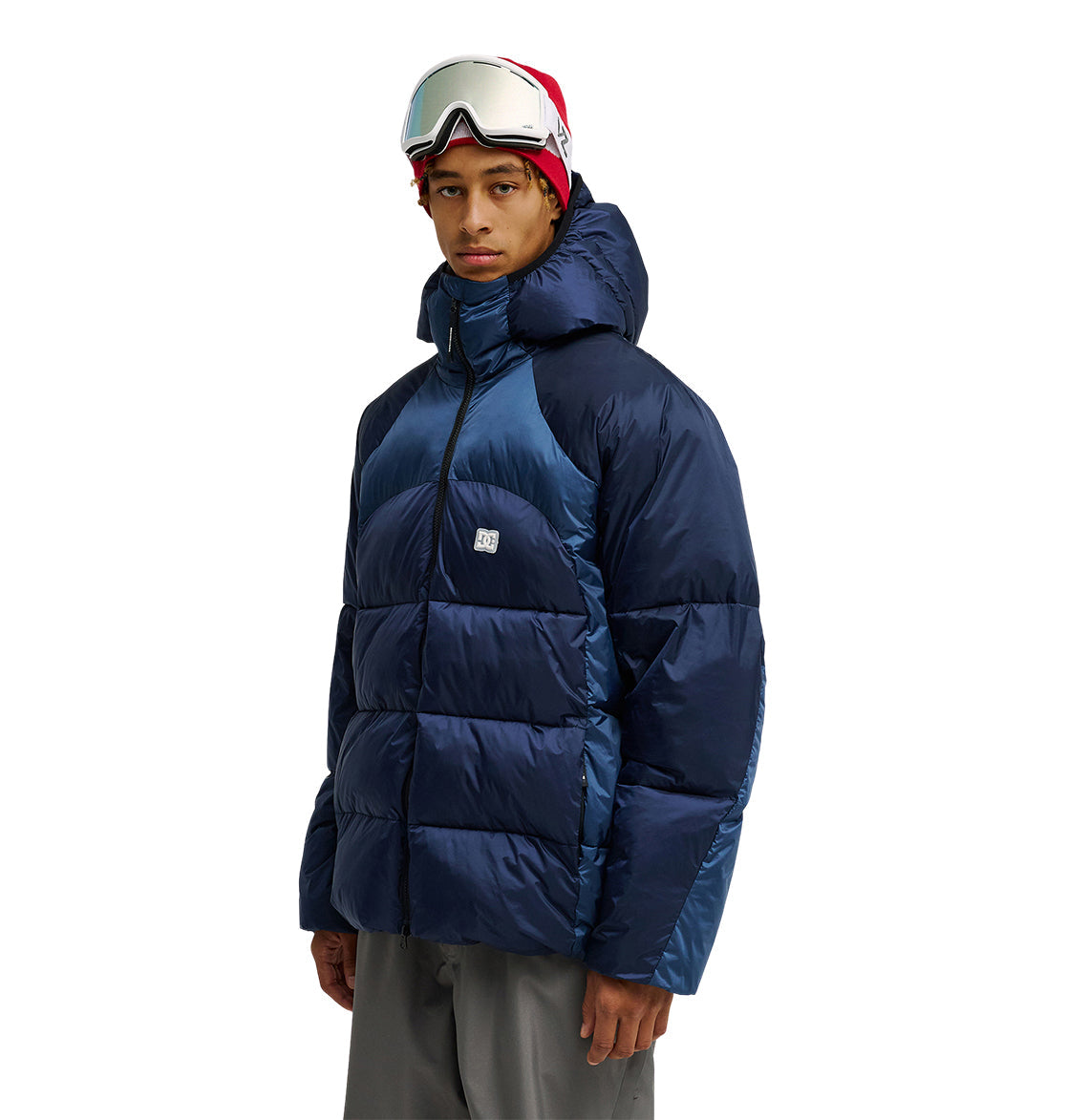 A person stands facing forward against a plain white background, wearing the DC Shoes Powder Puffer 10K jacket—renowned for its superior insulation and snow performance—styled with gray pants, a red beanie, and ski goggles.