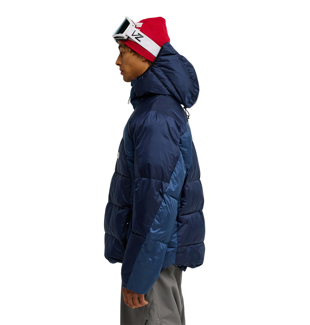 A person wearing the DC Shoes Powder Puffer 10K jacket in blue, paired with gray pants and a red beanie with ski goggles, stands in profile facing left against a white background, showcasing the Powder Puffer 10K’s superior insulation and snow performance.