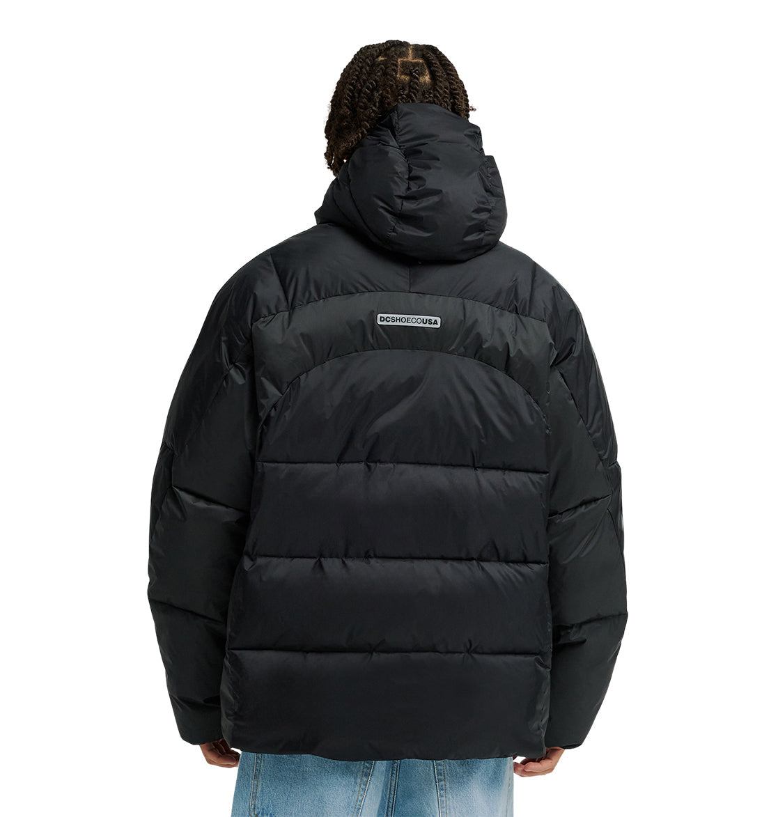 A person is seen from the back wearing the DC Shoes Powder Puffer 10K, a black hooded puffer jacket that provides breathable protection for all-day comfort. The jacket’s logo appears on the upper back. The outfit is completed with light blue jeans.