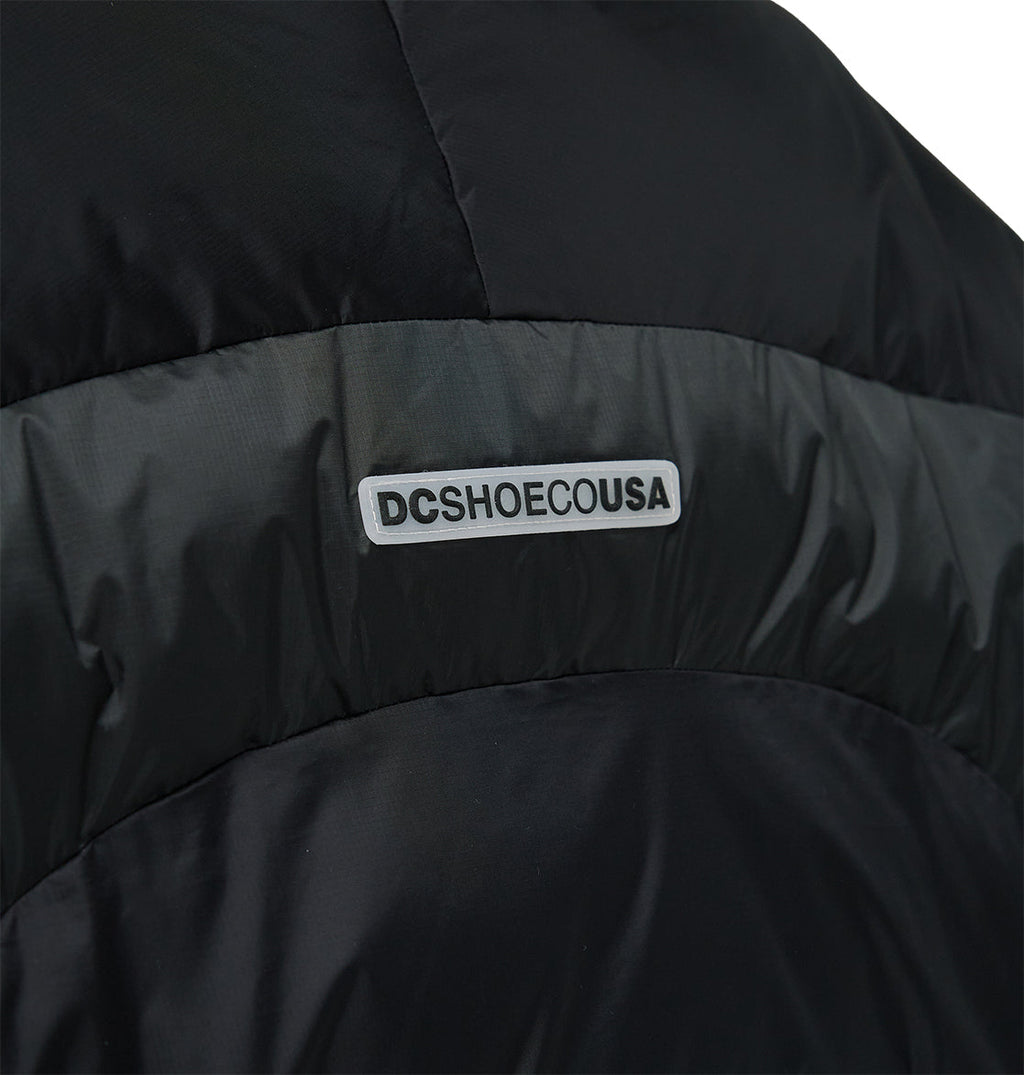 Close-up of the DC Shoes Powder Puffer 10K jacket in black, showcasing a horizontal reflective strip with “DCSHOECOUSA” in bold black letters on a white background. This jacket offers breathable protection for enhanced comfort and snow performance.