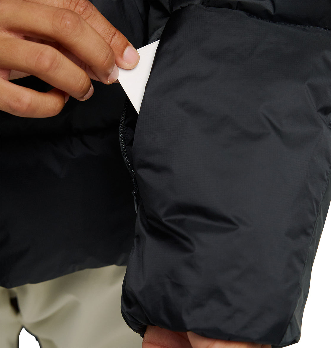 A hand slips a small white card into the zippered pocket of the DC Shoes Powder Puffer 10K jacket—a black puffer designed for optimal snow performance and breathable protection. The background is neutral, showing only the sleeve of the jacket and part of the pants.