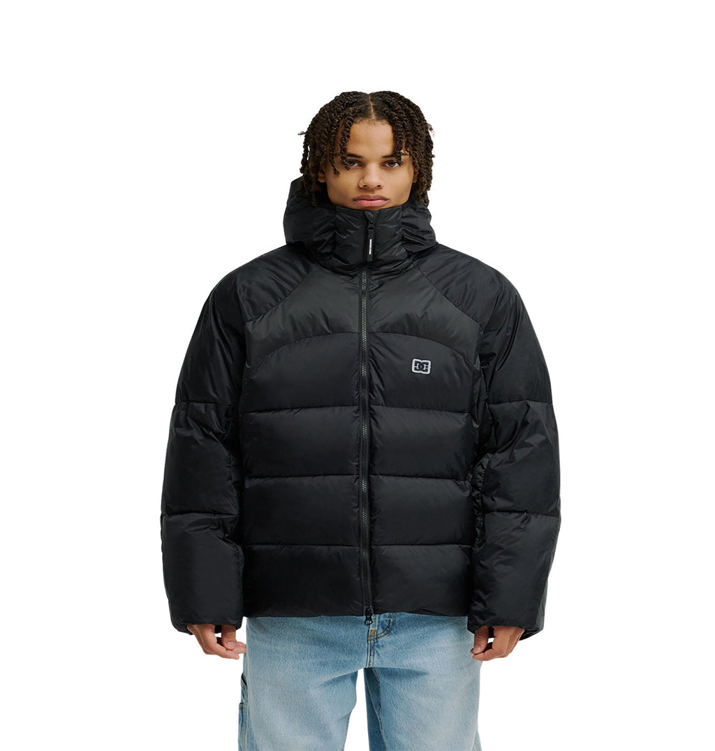 A person stands facing forward, wearing the DC Shoes Powder Puffer 10K—a black puffer jacket with a hood designed for breathable protection. The jacket features a small square logo on the chest and is zipped up. The individual has short, curly hair and is also wearing light blue jeans.