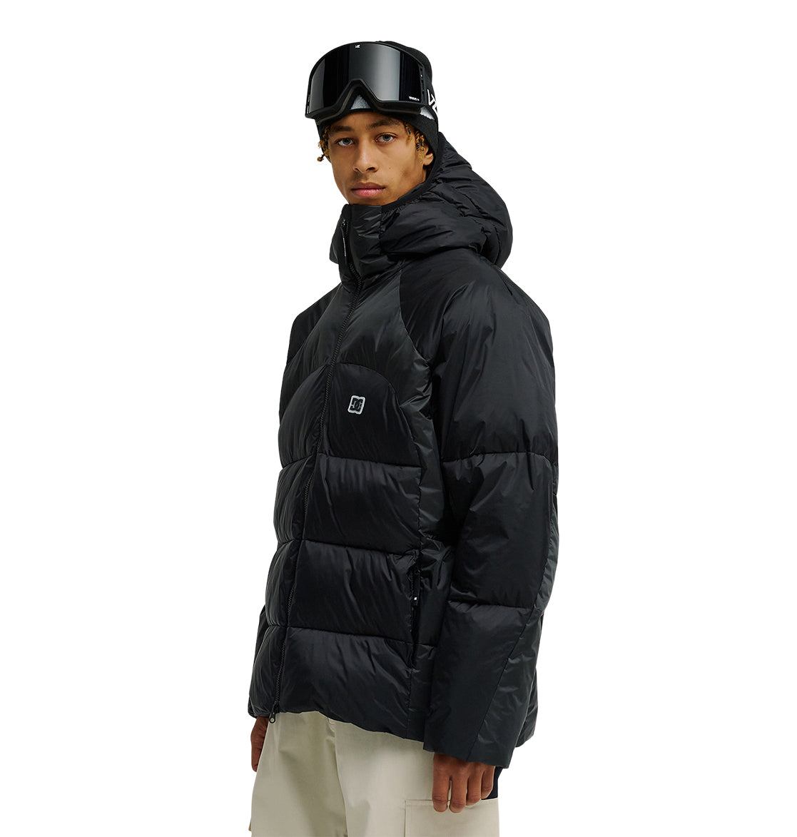 A person wearing the DC Shoes Powder Puffer 10K black jacket, paired with beige pants and black ski goggles, stands facing slightly to the side against a plain white background.
