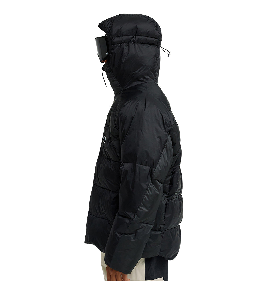 A person wearing the DC Shoes Powder Puffer 10K jacket with the hood up and oversized black goggles stands sideways against a plain white background, highlighting the jacket’s waterproofing and superior snow performance.