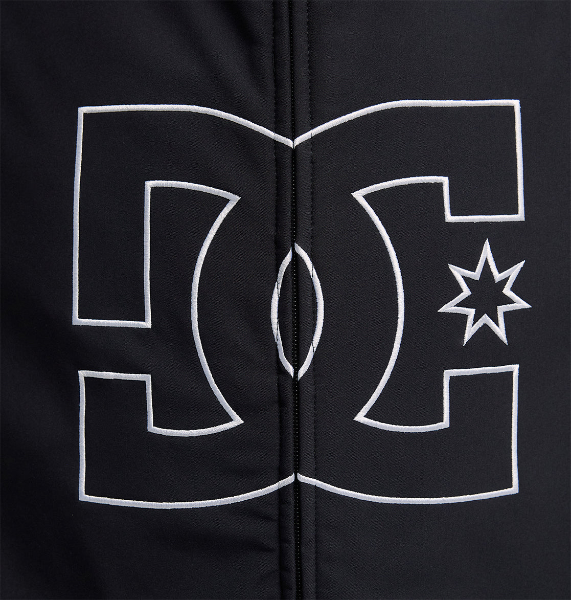 A prominent white DC Shoes logo, outlined in white, is centered on a black fabric background featuring visible vertical stitching—a design element characteristic of the Spectrum Softshell Jacket by DC Shoes, renowned for its waterproof and breathable protection.