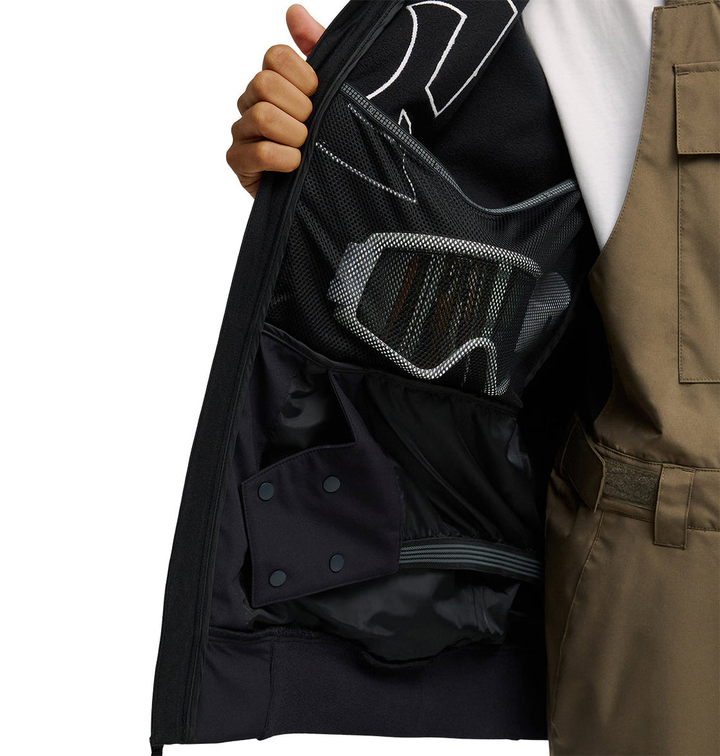 A person opens a DC Shoes Spectrum Softshell Jacket to reveal an interior mesh pocket holding ski goggles, while wearing tan overalls and a white shirt.
