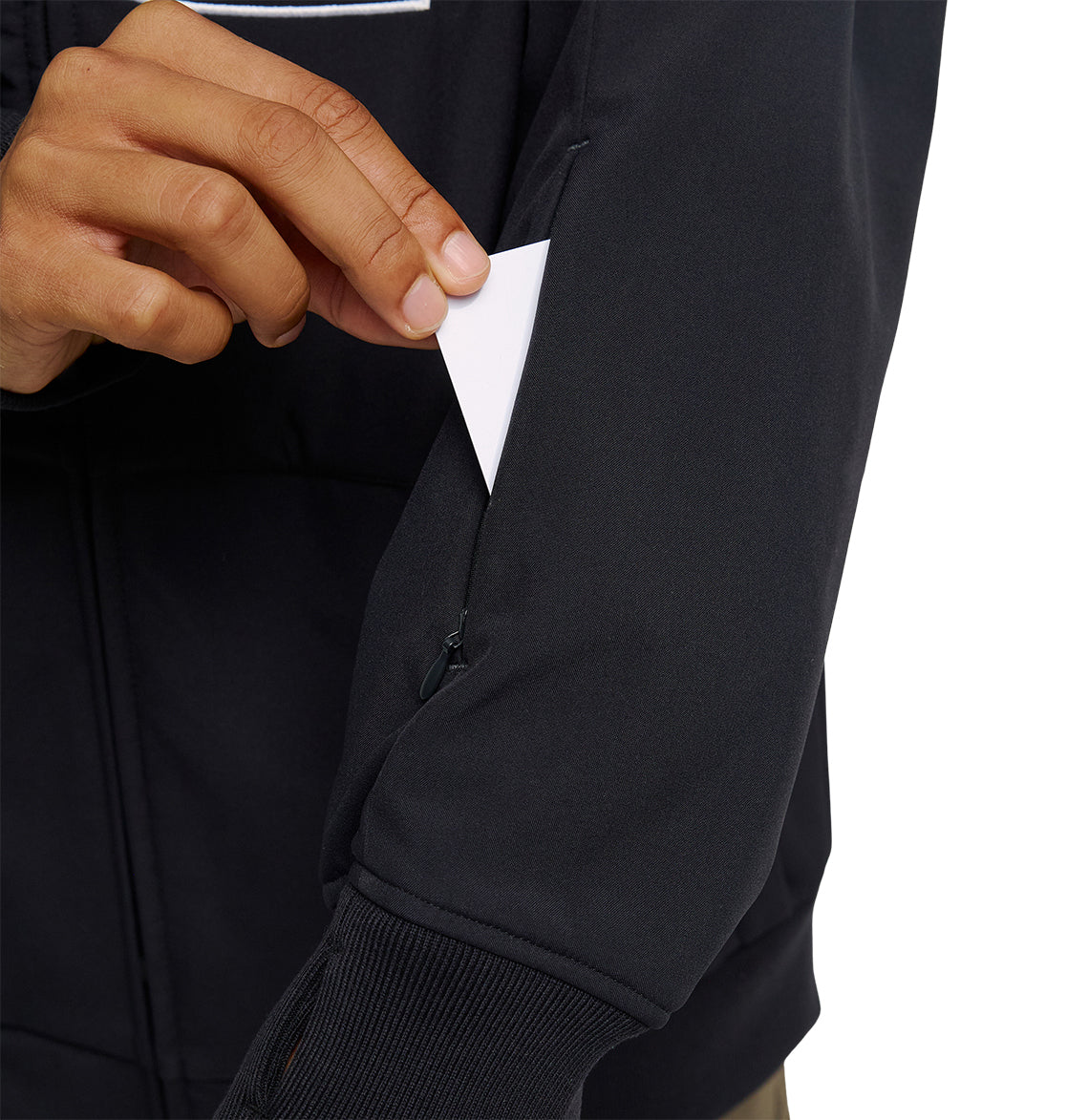 A person inserts a small white triangular card into a zippered pocket on the sleeve of the DC Shoes Spectrum Softshell Jacket, renowned for its excellent waterproofing and snow performance.