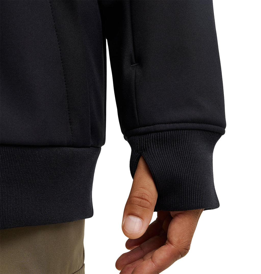A close-up of a person’s hand partially inside the sleeve of the DC Shoes Spectrum Softshell Jacket, highlighting a thumb hole designed for added comfort and breathable protection. The person is wearing light brown pants.