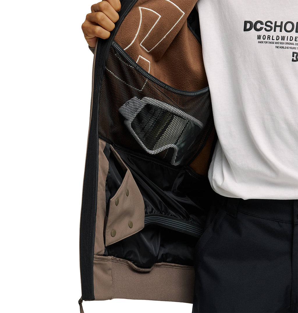 A person opens their DC Shoes Spectrum Softshell Jacket to reveal the inner pocket and mesh compartments, with ski goggles stored inside. The person is wearing a white DC Shoes Worldwide t-shirt and black pants.