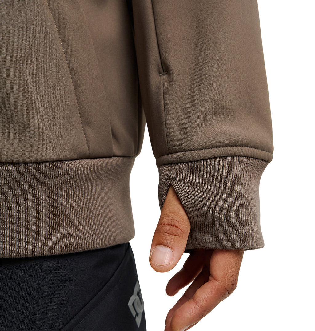 A close-up of a person wearing the DC Shoes Spectrum Softshell Jacket in brown, featuring a thumb hole in the sleeve cuff that reveals their thumb, paired with black pants.