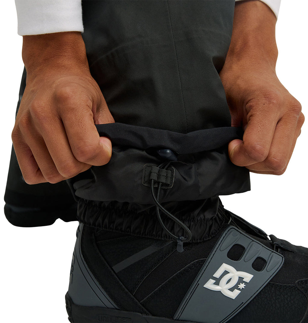 A person tightens the elastic cuff of their black, breathable Storm Division 3L Bib 30K snow pants from DC Shoes over a black DC snowboard boot, using a drawstring closure.
