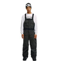 A person stands facing forward with hands at their sides against a plain white background, wearing the DC Shoes Storm Division 3L Bib 30K over dark attire, a white long-sleeve shirt with text, and a black beanie—showcasing this bib’s breathable protective design.