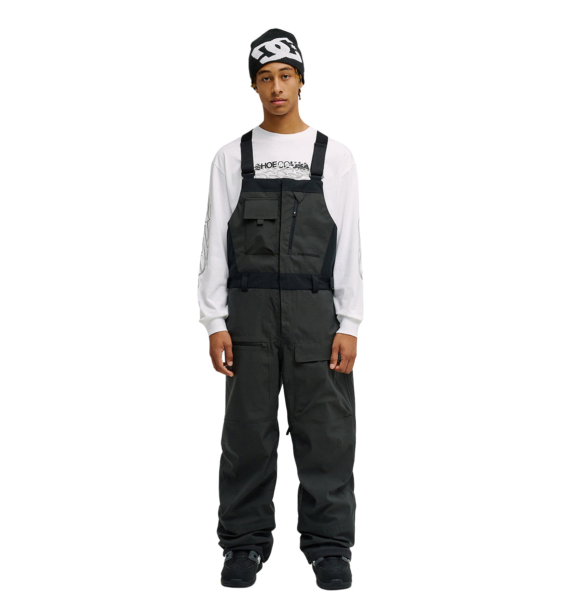A person stands facing forward with hands at their sides against a plain white background, wearing the DC Shoes Storm Division 3L Bib 30K over dark attire, a white long-sleeve shirt with text, and a black beanie—showcasing this bib’s breathable protective design.