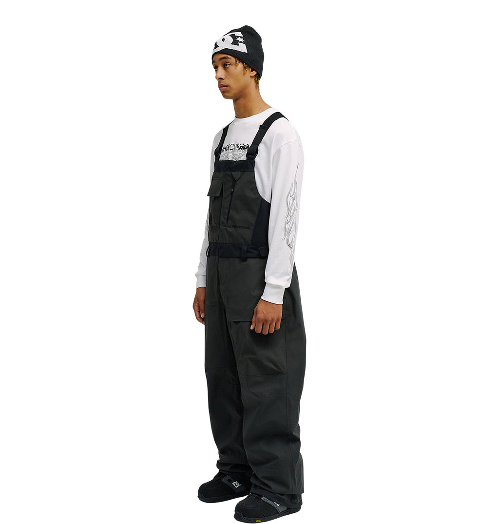 A person stands sideways against a white background wearing the DC Shoes Storm Division 3L Bib 30K in black, paired with a black beanie, a white graphic long-sleeve shirt, and black shoes—showcasing breathable protection engineered for enhanced snow performance.