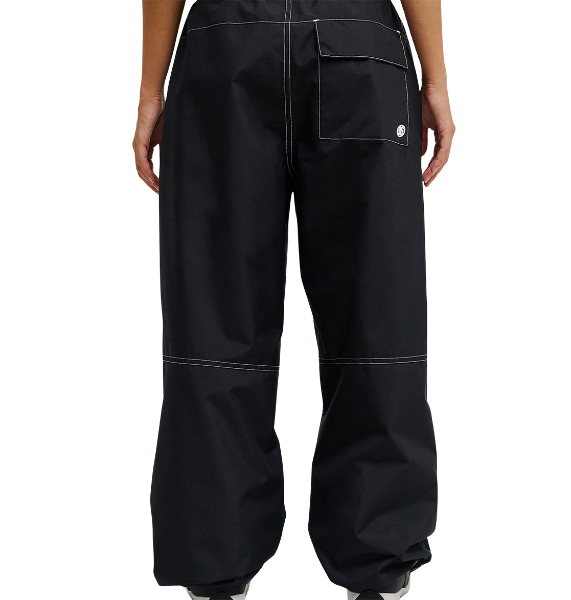 Person shown from the back wearing loose-fitting black DC Shoes Trackstar Pants 10K, featuring contrasting white stitching and a large right back pocket. The hems drape over most of their shoes. Designed for snow performance, these pants offer waterproofing details. Hands rest at their sides.