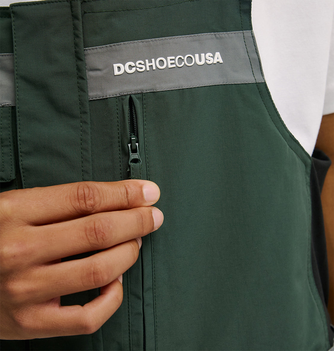 A hand unzips the DC Shoes Mountain Lab Bib 20K in green and gray, which features “DCSHOECOUSA” printed on the chest and offers breathable protection. The person is wearing a white shirt underneath.