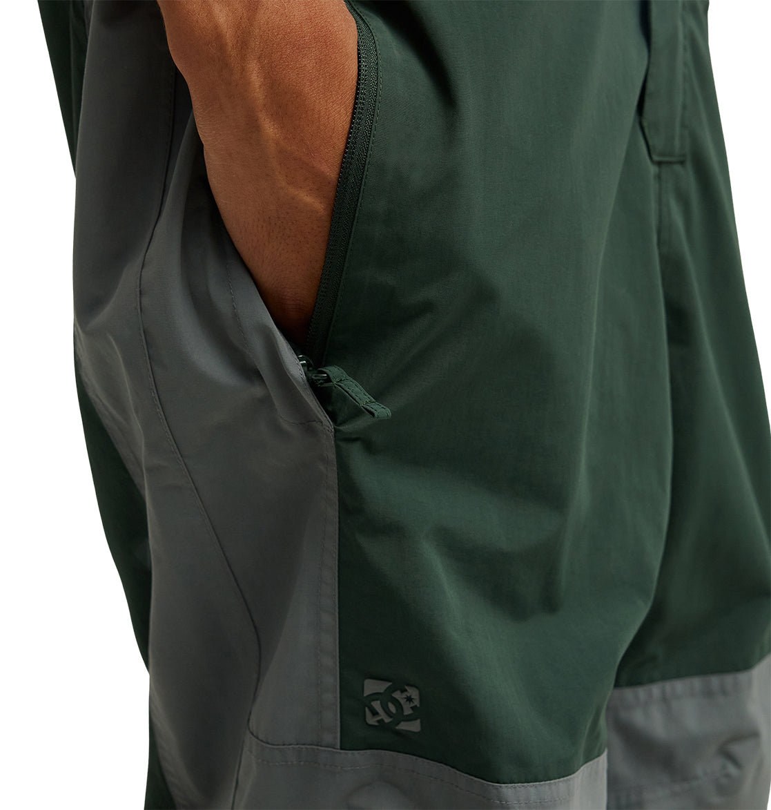 Close-up of a person’s hand in the zipper pocket of dark green and gray DC Shoes Mountain Lab Bib 20K, featuring breathable protection for added comfort and the DC Shoes logo near the hem.