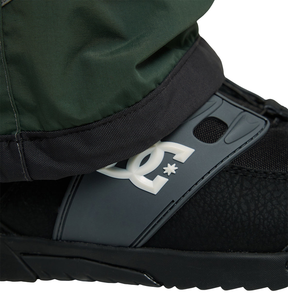 A close-up of a person’s pant leg from the DC Shoes Mountain Lab Bib 20K, an insulated snow bib, tucked into a black snowboard boot with a gray strap featuring a prominent white DC Shoes logo.