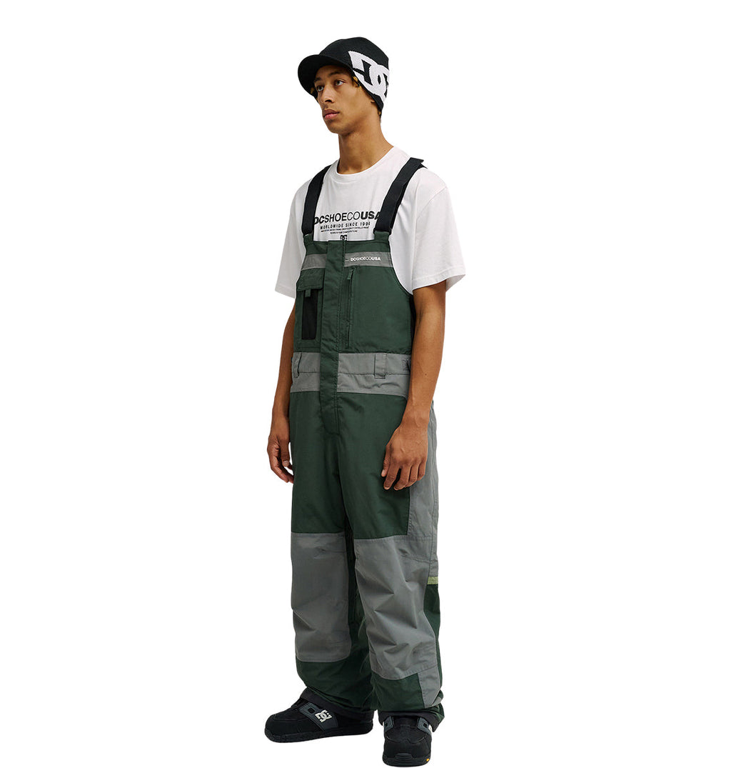 A person wearing a black and white beanie, a white T-shirt, and the green and gray DC Shoes Mountain Lab Bib 20K stands facing left with a neutral expression against a plain white background.