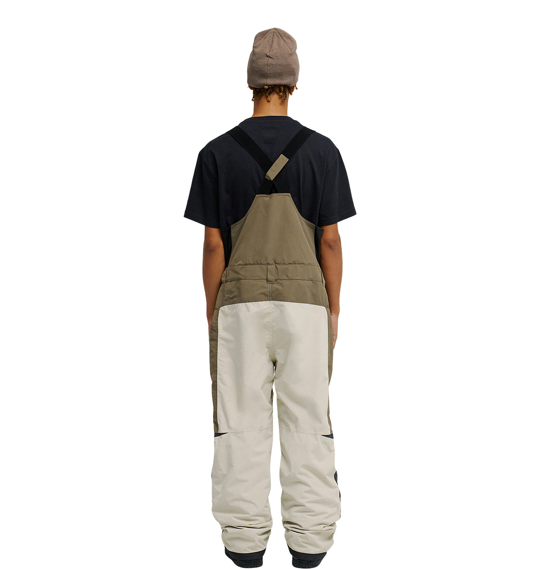 A person wearing a black T-shirt, the DC Shoes Mountain Lab Bib 20K two-tone overalls (brown top, beige bottom) designed for breathable protection, and a beige beanie stands facing away from the camera against a white background.