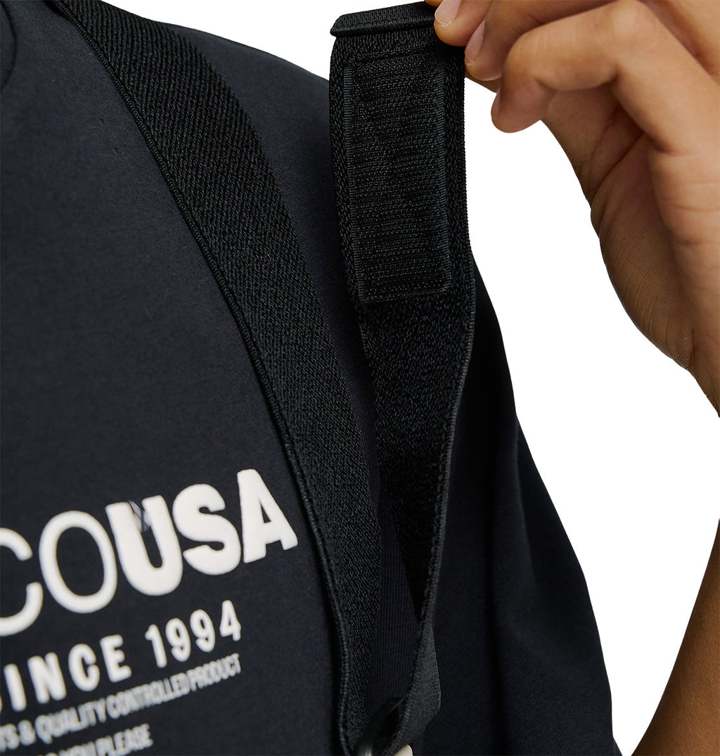 Wearing a dark shirt with white text partially reading “COUSA SINCE 1994,” a person adjusts the black strap of the Mountain Lab Bib 20K from DC Shoes, ready for any adventure with breathable protection.