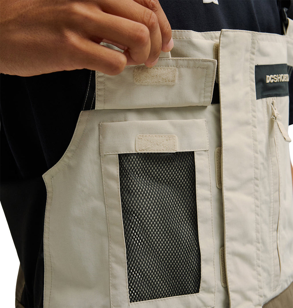 A person lifts the flap of a beige Mountain Lab Bib 20K pocket with mesh detailing by DC Shoes, revealing a Velcro closure. The bib, designed for breathable protection, is worn over a dark shirt.