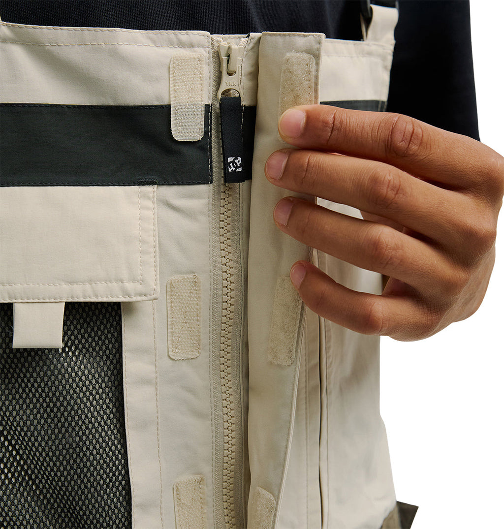 A person uses their right hand to zip up the DC Shoes Mountain Lab Bib 20K, appreciating its beige color, Velcro patches, mesh pocket, and breathable protection.