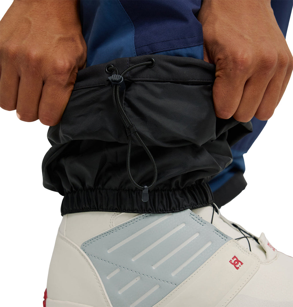 Close-up of hands adjusting the drawstring on the cuff of DC Shoes’ Mountain Lab Pants 10K in black, worn over a white and light blue sneaker with red accents, showcasing their breathable protection for enhanced comfort and performance.